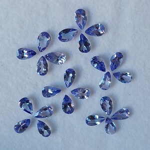 May include: A collection of pear-shaped gemstones in various shades of blue, arranged in a loose pattern on a white surface.