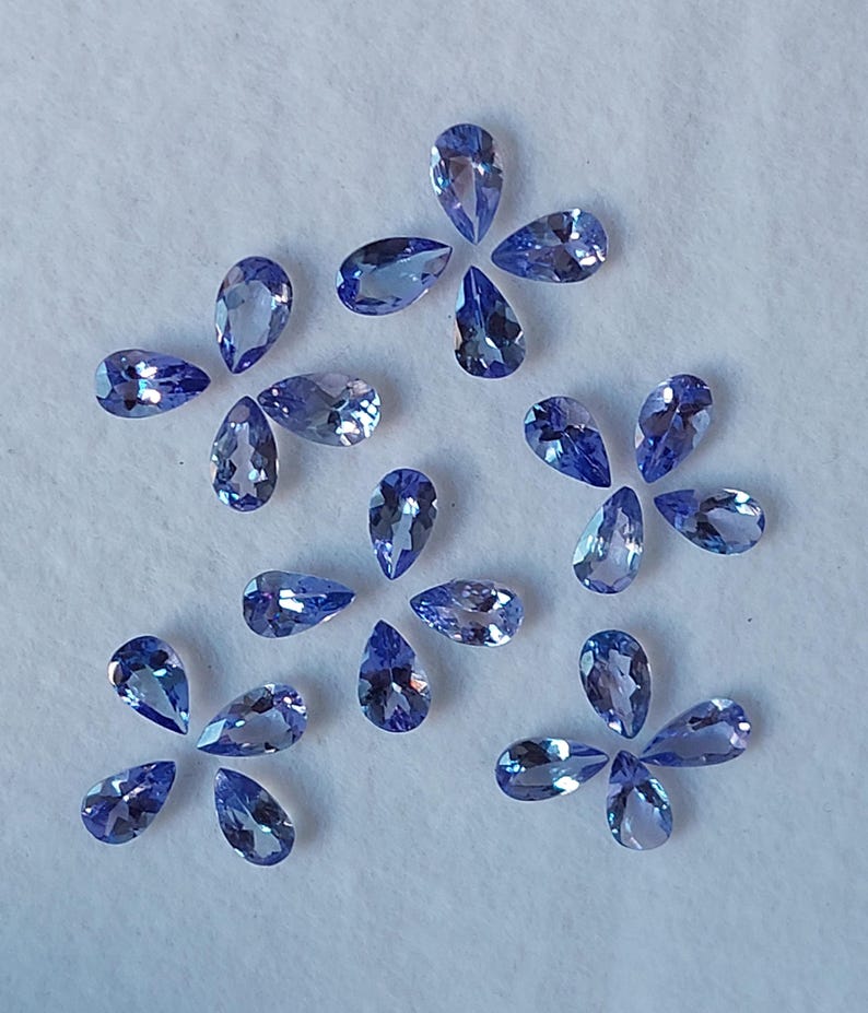May include: A collection of pear-shaped gemstones in various shades of blue, arranged in a loose pattern on a white surface.