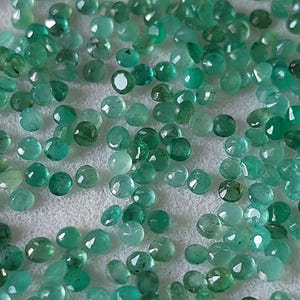 May include: A scattering of small, round emerald gemstones in various shades of green. The gemstones are spread across a white surface, with facets reflecting light. The gems are of different sizes and shapes.