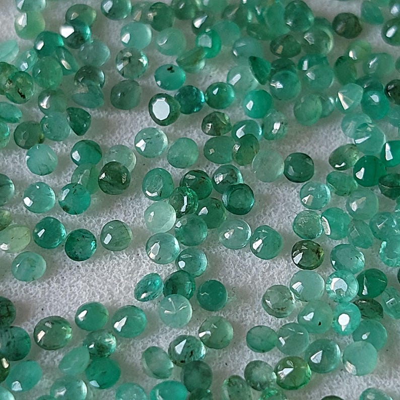 May include: A scattering of small, round emerald gemstones in various shades of green. The gemstones are spread across a white surface, with facets reflecting light. The gems are of different sizes and shapes.