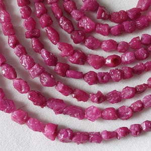 May include: Strands of rough-cut ruby gemstones in varying shapes and sizes. The gemstones are a deep, vibrant pinkish-red color. The beads are strung together, creating a layered effect.