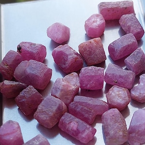 Może przedstawiać: Kolekcja surowych, nieoszlifowanych kamieni szlachetnych rubinu o różnych kształtach i rozmiarach. Rubiny wykazują odcienie różu i czerwonoróżowego, z szorstkimi, teksturowanymi powierzchniami. Kamienie szlachetne są rozrzucone na jasnej powierzchni.