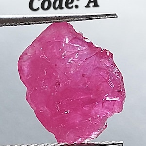 May include: A pink rough ruby gemstone held in tweezers. The gemstone is a deep pink color and has a rough, natural texture. Code: A