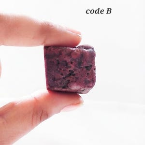以下が含まれることがあります： 濃い紫色のテクスチャーのある多孔質な表面を持つ、小さな立方体に近い形状の物体。右上に「code B」というテキストが記されています。このアイテムは、暗い斑点や色の変化が見られる不規則で自然な外観をしています。