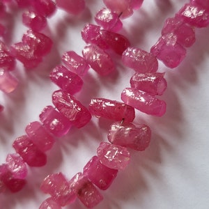 May include: A strand of pink ruby beads, each bead is faceted and has a rough, irregular shape. The beads are strung on a clear thread and are arranged in a loose, flowing pattern.