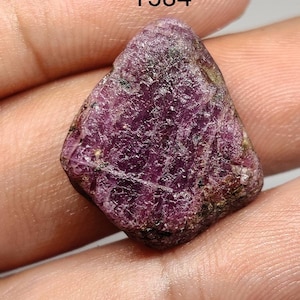 Puede incluir: Una gema de color púrpura oscuro y áspera con una superficie rugosa y desigual. La gema se está sosteniendo en una mano. El número 1584 es visible en la imagen.