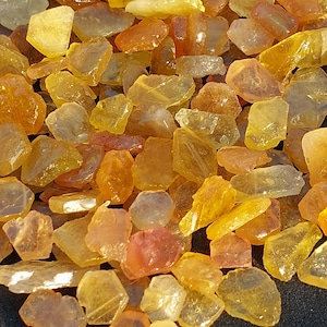 Pode incluir: Uma coleção de pequenos pedaços irregulares de âmbar em vários tons de amarelo, laranja e marrom. As peças de âmbar são translúcidas e têm um acabamento brilhante.