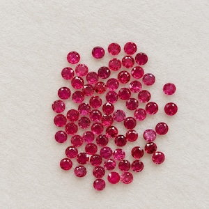 May include: A collection of numerous small, round, faceted loose gemstones in a vibrant, deep red color. Each stone features a brilliant cut with multiple facets that reflect light, creating a sparkling appearance across the group.