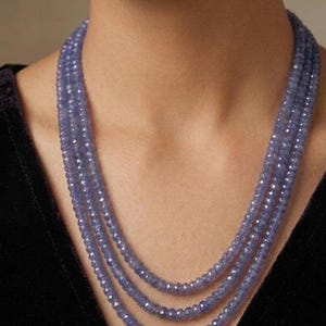 Puede incluir: Un collar de tres hilos compuesto por pequeñas cuentas facetadas de color azul bígaro. Las cuentas son uniformes en tamaño y forma, creando un efecto de capas alrededor del cuello. El collar presenta una textura y color constantes en cada hilo.