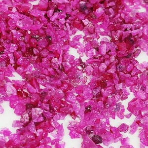 May include: A close-up view of a large quantity of small, pink ruby gemstones. The rubies are scattered on a white surface.