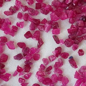 May include: A close-up view of a collection of small, rough, pink ruby gemstones. The rubies are scattered on a white surface.
