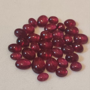 May include: A collection of oval-shaped, cabochon cut, red gemstones. The gemstones are various sizes and are arranged in a loose circle on a white surface.