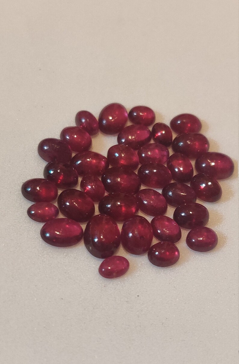 May include: A collection of oval-shaped, cabochon cut, red gemstones. The gemstones are various sizes and are arranged in a loose circle on a white surface.
