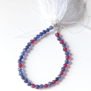 Puede incluir: Una hilera de cuentas redondas facetadas en colores azul profundo y rojo translúcido, dispuestas en un bucle circular. Las cuentas están ensartadas en un hilo fino que se reúne en la parte superior y termina con una larga borla decorativa blanca.