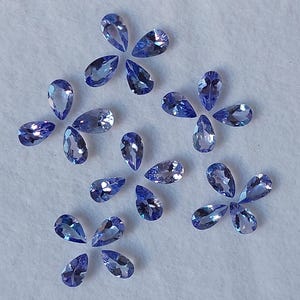 May include: A collection of pear-shaped, faceted gemstones in various shades of blue, arranged on a white surface.