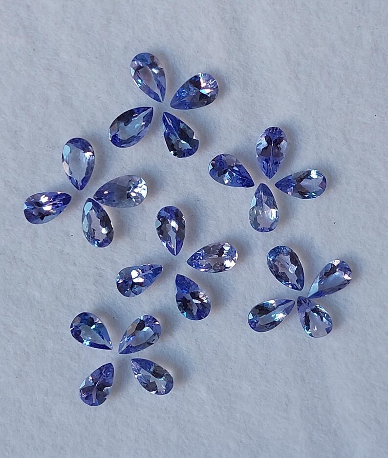 May include: A collection of pear-shaped, faceted gemstones in various shades of blue, arranged on a white surface.