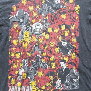 Op de afbeelding: Zwart t-shirt met een collage van Iron Man-pakontwerpen in rood, geel, grijs en zwart. De afbeelding bevat verschillende versies van het Iron Man-pantser, met het woord "IRON MAN" in het wit onderaan gedrukt.