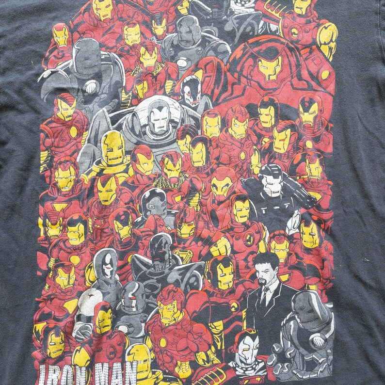 Op de afbeelding: Zwart t-shirt met een collage van Iron Man-pakontwerpen in rood, geel, grijs en zwart. De afbeelding bevat verschillende versies van het Iron Man-pantser, met het woord "IRON MAN" in het wit onderaan gedrukt.