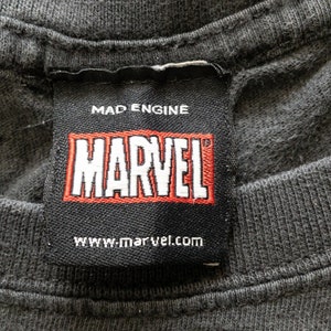 Op de afbeelding: Close-up van een zwart t-shirtlabel met het MARVEL-logo in rood en wit. Het label bevat ook de tekst "MAD ENGINE" en "www.marvel.com". De stof van het t-shirt is donkergrijs.