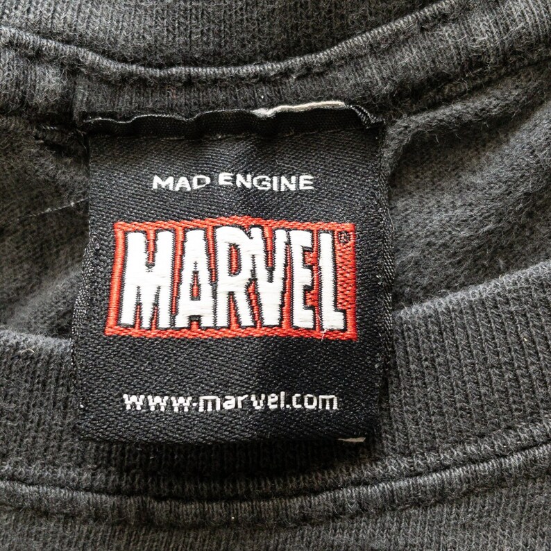 Op de afbeelding: Close-up van een zwart t-shirtlabel met het MARVEL-logo in rood en wit. Het label bevat ook de tekst "MAD ENGINE" en "www.marvel.com". De stof van het t-shirt is donkergrijs.