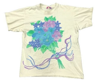Puede incluir: Camiseta color crema con un diseño de ramo floral. El ramo incluye flores azules, moradas y rosas con hojas verdes. Cintas azules y moradas acentúan el diseño. La camiseta tiene mangas cortas y cuello redondo.