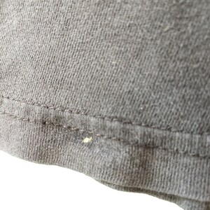 Op de afbeelding: Close-up van een deel van een donkergrijs t-shirt. De stof heeft een getextureerde uitstraling met zichtbare stiksels langs de zoom. Er is een kleine, lichtgekleurde vlek aanwezig op de stof.
