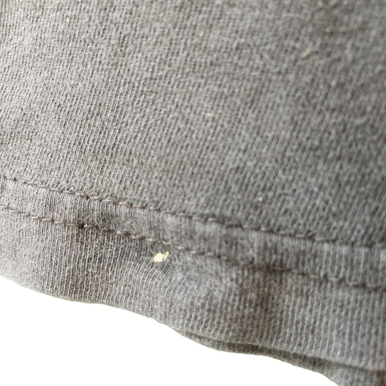 Op de afbeelding: Close-up van een deel van een donkergrijs t-shirt. De stof heeft een getextureerde uitstraling met zichtbare stiksels langs de zoom. Er is een kleine, lichtgekleurde vlek aanwezig op de stof.