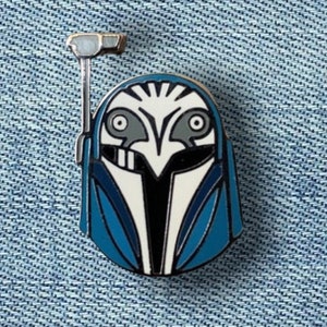 May include: Enamel pin featuring a stylized blue, white, and black helmet design with a silver-colored attachment. The pin has a detailed, symmetrical design, with a blue outline and a white and black face. The pin is set against a denim background.