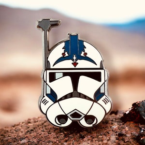 May include: A white and blue enamel pin featuring a Star Wars clone trooper helmet with a blue and white design. The helmet has a blue and white crest on the top and a black visor.