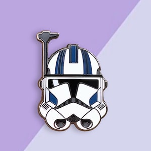 May include: A white and blue enamel pin featuring a Star Wars clone trooper helmet. The helmet has a black visor and a blue and white stripe pattern.