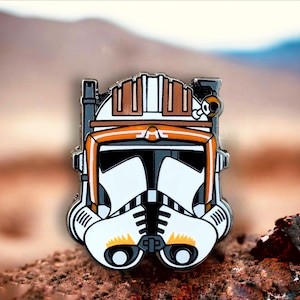 May include: A white and orange enamel pin featuring a cartoon depiction of a Star Wars clone trooper helmet. The helmet has a white background with orange accents and a black visor.