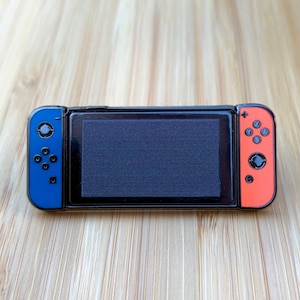 Op de afbeelding: Een zwarte emaille pin in de vorm van een Nintendo Switch-console. De linkerzijde van de console is blauw met zwarte knoppen, en de rechterzijde is oranje met zwarte knoppen. Het scherm is zwart.