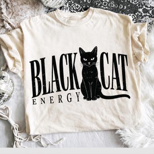 May include: A cream-colored short-sleeve t-shirt featuring a black graphic of a sitting cat. The text "BLACK CAT" is printed in a large, distressed black font, with the word "ENERGY" positioned below in a smaller, matching black font.