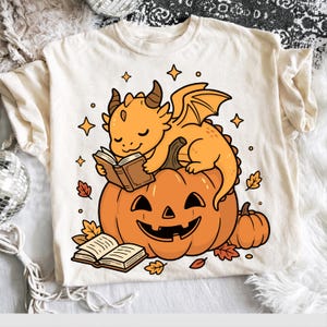 May include: A cream-colored t-shirt featuring a graphic illustration of an orange dragon sitting on a large jack-o'-lantern while reading a book. Another open book lies in front of the pumpkin, surrounded by small orange autumn leaves and stars.