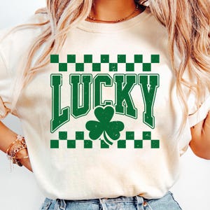 May include: Cream-colored t-shirt with a green "LUCKY" graphic and a shamrock. The word "LUCKY" is in a bold, distressed font, with a green and white checkered pattern above and below the text. The shamrock is centered below the word.