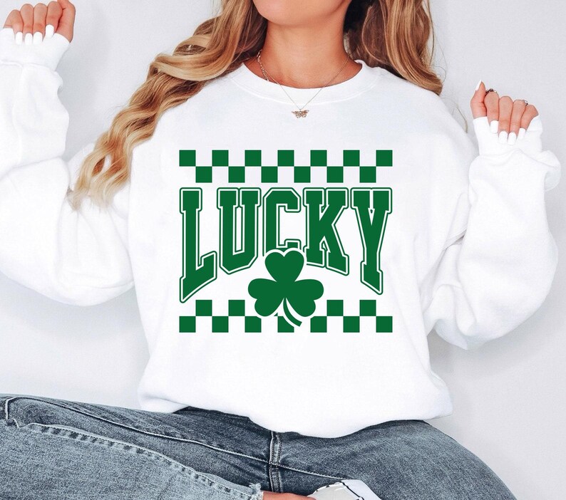 May include: White sweatshirt with the word "LUCKY" in green block letters, a green shamrock, and a green and black checkered pattern. The sweatshirt is a casual style.