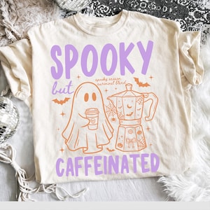Puede incluir: Una camiseta de color crema con el texto morado SPOOKY but CAFFEINATED. El gráfico incluye un fantasma naranja de líneas que sostiene una taza de café, una cafetera moka, murciélagos, telarañas y el texto spooky season survival blend en letra pequeña.