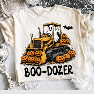 May include: A cream-colored t-shirt featuring a graphic of a yellow bulldozer driven by a white ghost. The bulldozer is loaded with orange jack-o'-lantern pumpkins, and a black bat flies above. The text BOO-DOZER is printed in black below.