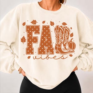 May include: A cream-colored crewneck sweatshirt featuring a graphic print with the text FALL vibes in orange. The letters F, A, and L are decorated with white polka dots, while the letter L is replaced by a pair of orange cowboy boots and a pumpkin.