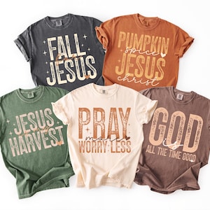 May include: Five short-sleeve crew neck t-shirts in charcoal, orange, olive green, cream, and brown. Each shirt features graphic text designs with religious themes, including phrases like Fall for Jesus, Pumpkin Spice Jesus, Jesus Harvest, Pray More Worry Less, and God is All the Time Good.