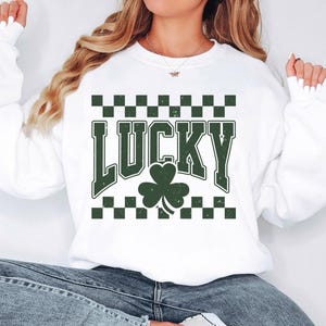 May include: White sweatshirt with the word "LUCKY" in green block letters, a green shamrock, and a green and black checkered pattern. The sweatshirt is a casual style, perfect for St. Patrick's Day.
