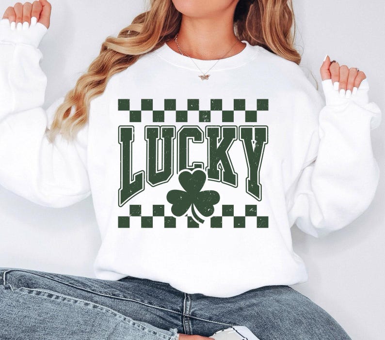 May include: White sweatshirt with the word "LUCKY" in green block letters, a green shamrock, and a green and black checkered pattern. The sweatshirt is a casual style, perfect for St. Patrick's Day.