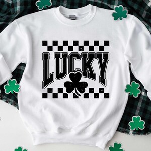 May include: White sweatshirt with the word "LUCKY" in black, distressed font, and a black shamrock. The word is set against a black and white checkered pattern. Green shamrock decorations are scattered around the sweatshirt.