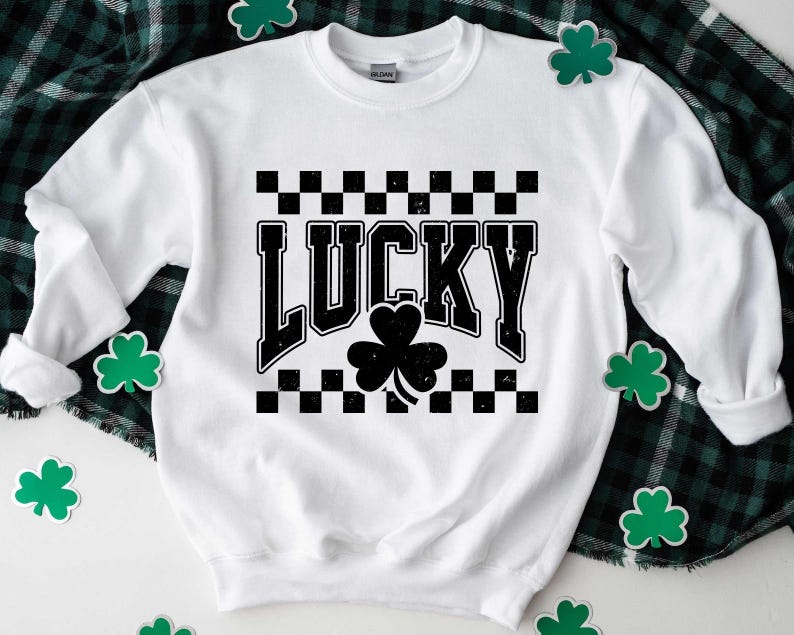 May include: White sweatshirt with the word "LUCKY" in black, distressed font, and a black shamrock. The word is set against a black and white checkered pattern. Green shamrock decorations are scattered around the sweatshirt.