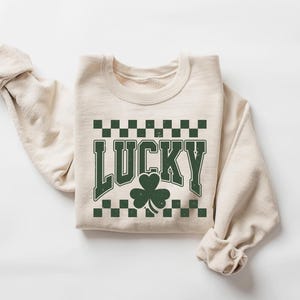 May include: A cream-colored sweatshirt with the word "LUCKY" in distressed green lettering, a green shamrock, and a green and black checkered pattern. The sweatshirt is folded with the sleeves rolled up.