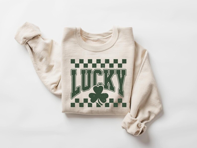 May include: A cream-colored sweatshirt with the word "LUCKY" in distressed green lettering, a green shamrock, and a green and black checkered pattern. The sweatshirt is folded with the sleeves rolled up.
