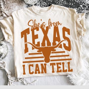 Op de afbeelding: Een crèmekleurig T-shirt met korte mouwen en tekst in gebrand oranje met de tekst She's from TEXAS I CAN TELL. Het ontwerp bevat een afbeelding van een silhouet van een longhorn-rund tussen het woord TEXAS en drie horizontale lijnen.