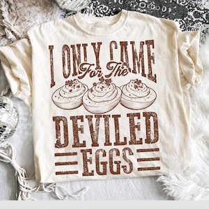 May include: A cream-colored short-sleeve t-shirt featuring a brown graphic print of three deviled eggs and the text "I ONLY CAME For The DEVILED EGGS" in a distressed, vintage-style font. The design is centered on the front of the garment.