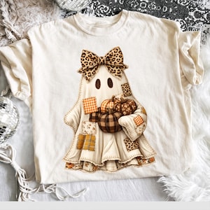 May include: A cream-colored t-shirt featuring a central graphic of a ghost wearing a leopard print bow. The ghost is decorated with various patches in plaid and leopard patterns and is holding a cluster of three pumpkins in brown and orange tones.