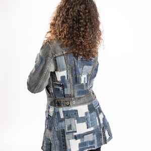 May include: A long-sleeved denim jacket constructed from various shades of blue and white fabric patches. The garment features a fitted waist with a horizontal band, two metal buttons at the back, and a flared hemline with raw-edged seams.